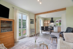 A spacious living area with a television, a dining table with chairs, a kitchen in the background, and large windows offering a view of the outdoors.
