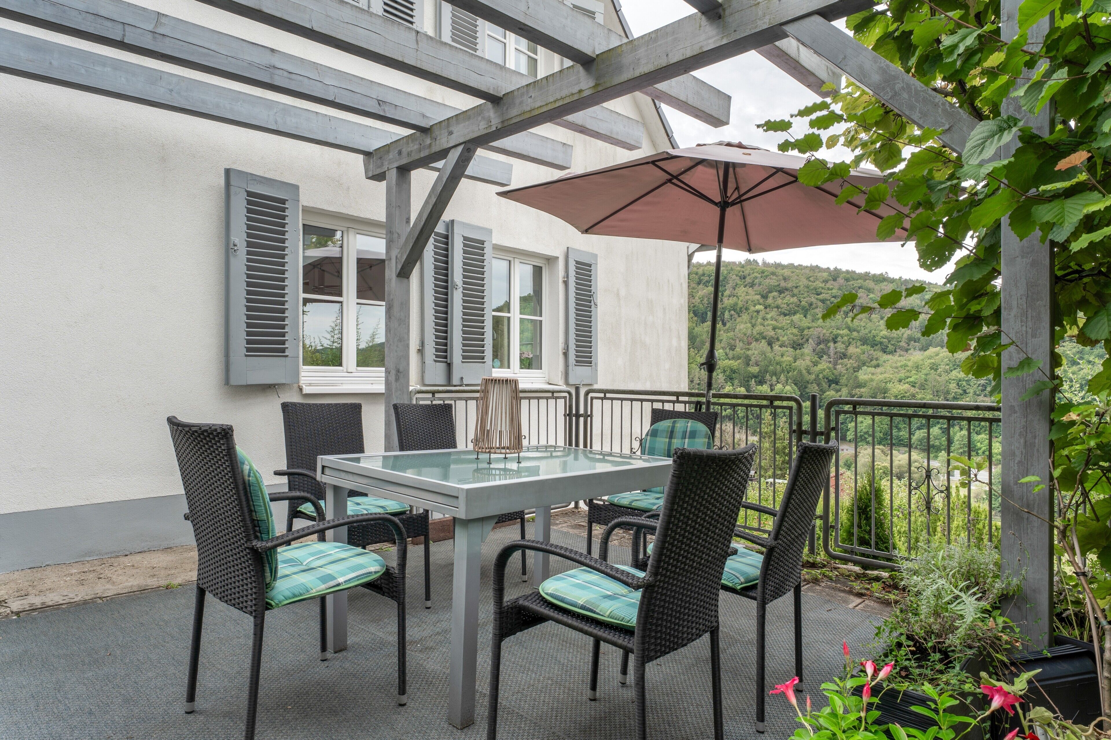 Un patio al aire libre con una mesa, sillas, un pérgola y vistas a una ladera boscosa.