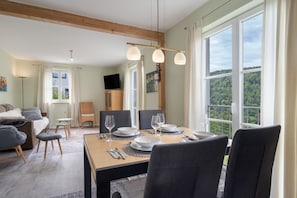 Un comedor con una mesa dispuesta para cuatro, un televisor al fondo y vistas a un paisaje boscoso a través de amplios ventanales.