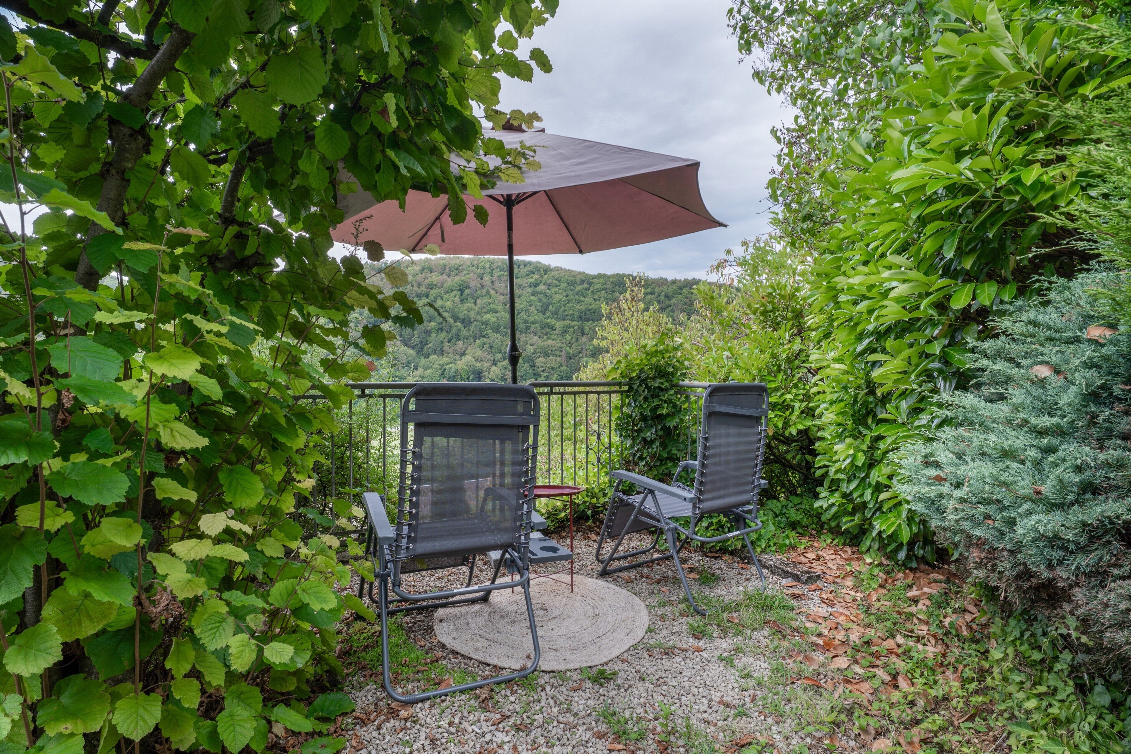 An outdoor seating area with two chairs and a table, shaded by a large umbrella, surrounded by lush greenery.