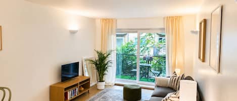 Una sala de estar moderna con un sofá, un televisor sobre un mueble de madera, una alfombra estampada y un balcón con plantas.