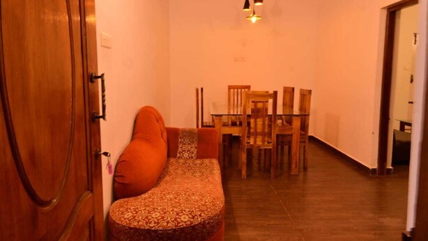 Un pasillo estrecho con una puerta de madera a la izquierda, un sofá con cojines estampados y un comedor con sillas y una mesa de madera.