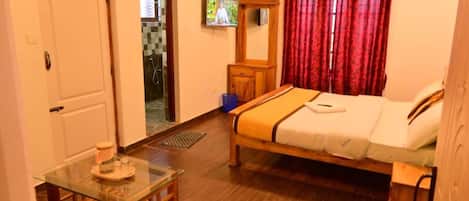 Habitación de hotel con cama, una mesa de vidrio, un armario de madera, un televisor y una puerta que da a un baño.