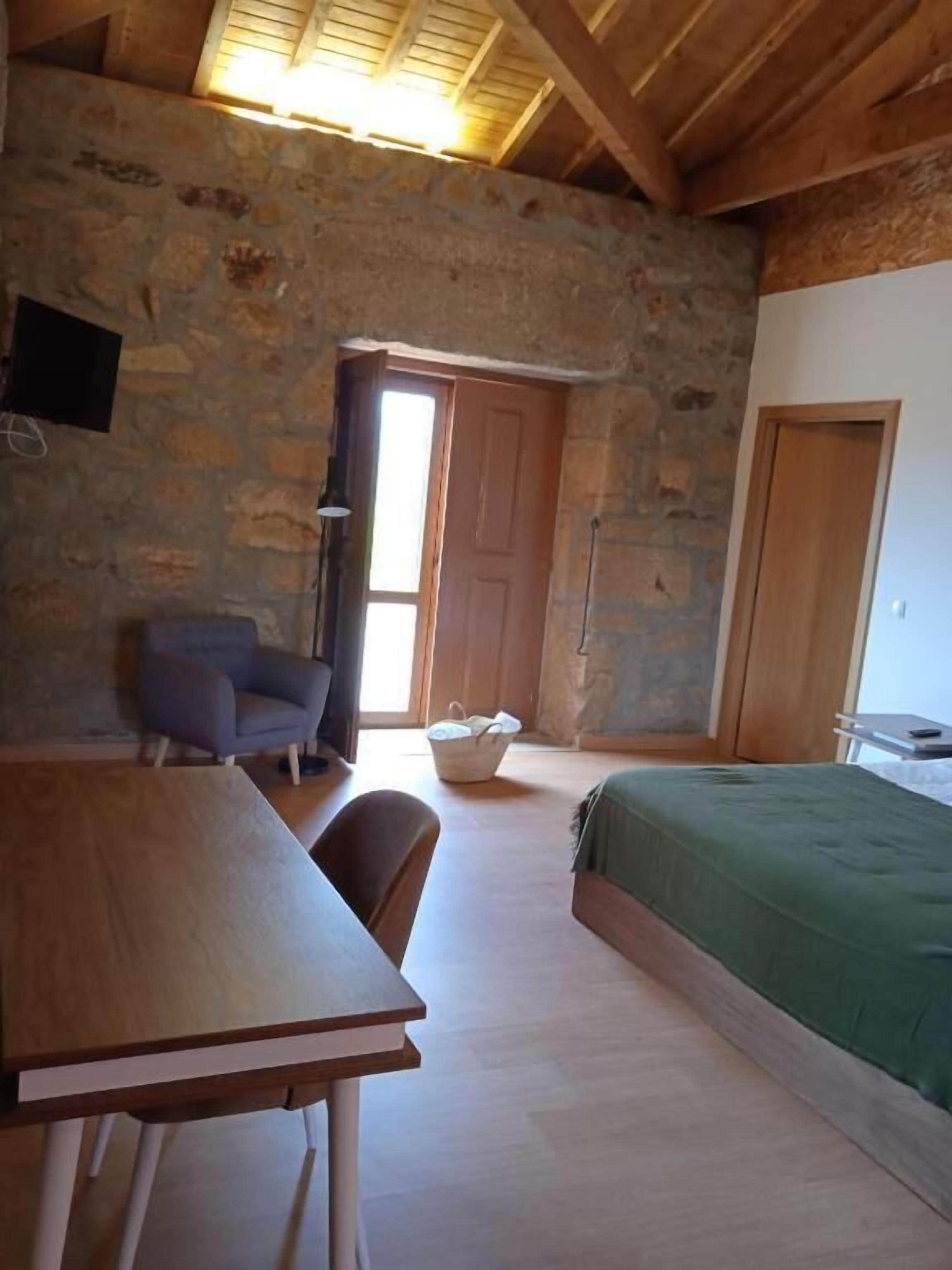 A room with a wooden table, a chair, a bed with a green cover, and a stone wall.