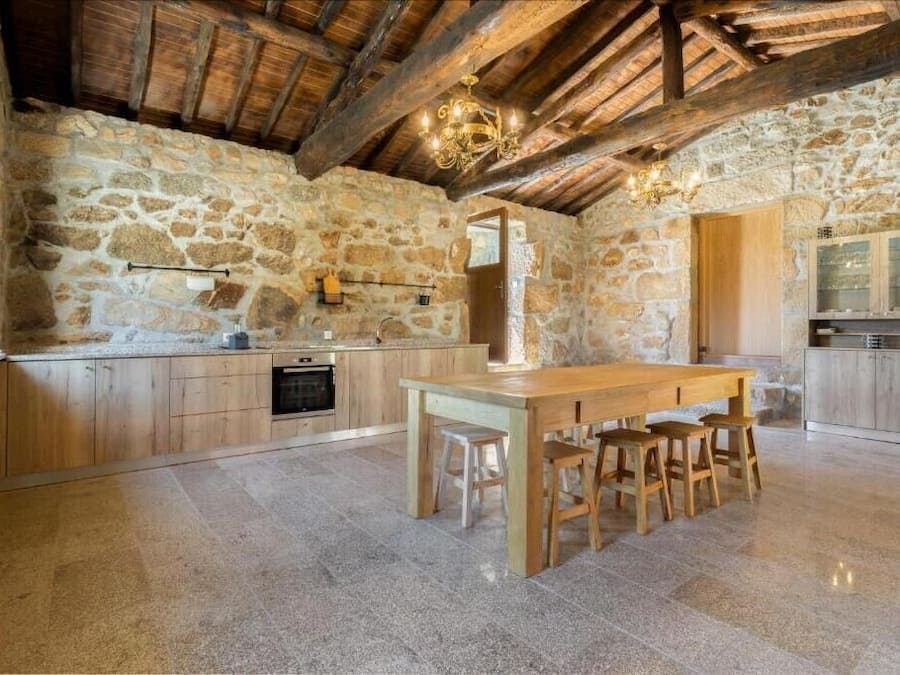A rustic kitchen with stone walls, wooden beams, and a wooden dining table with stools.