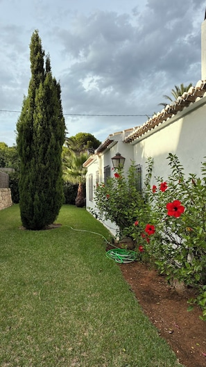 A well-maintained garden with a tall coniferous tree, a white building with a tiled roof, and red flowering plants. - A home with a Mediterranean feel in one of Jávea's most exclusive residential developments (Jávea)