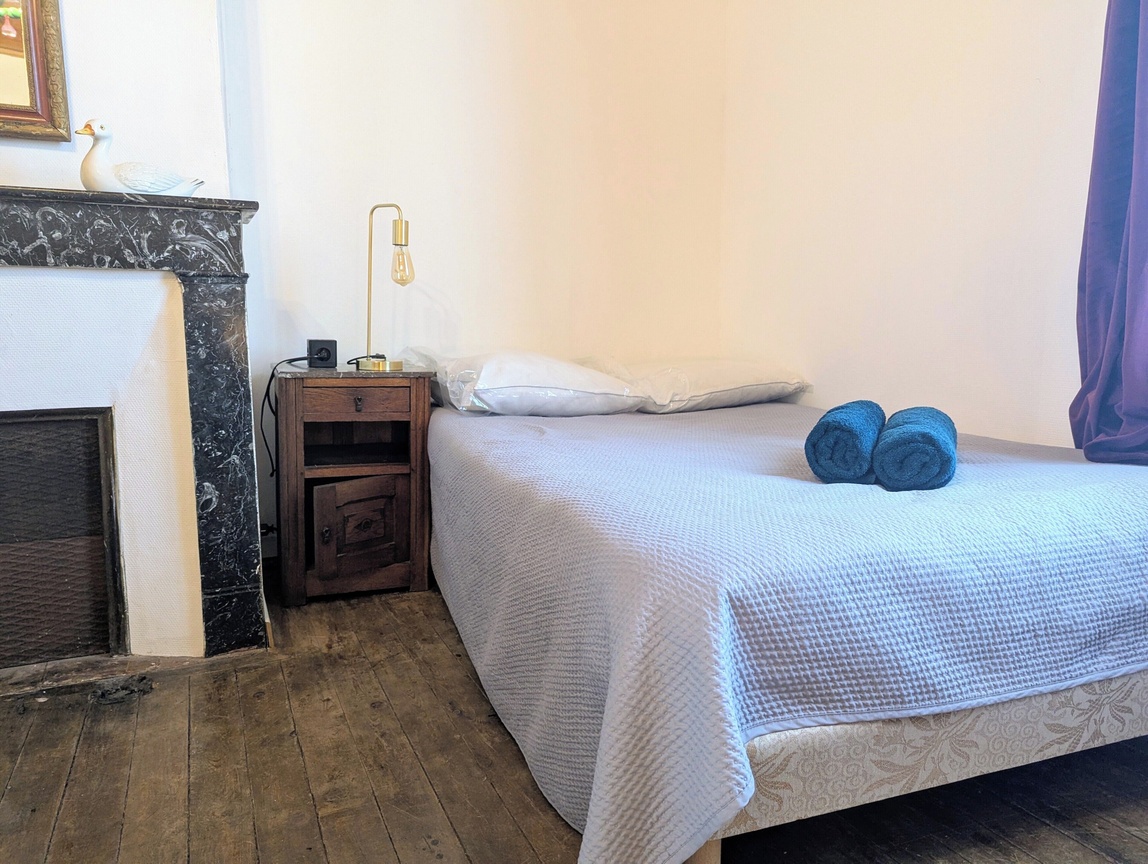 A bedroom with a bed, a nightstand, a lamp, a fireplace, and a window with curtains.