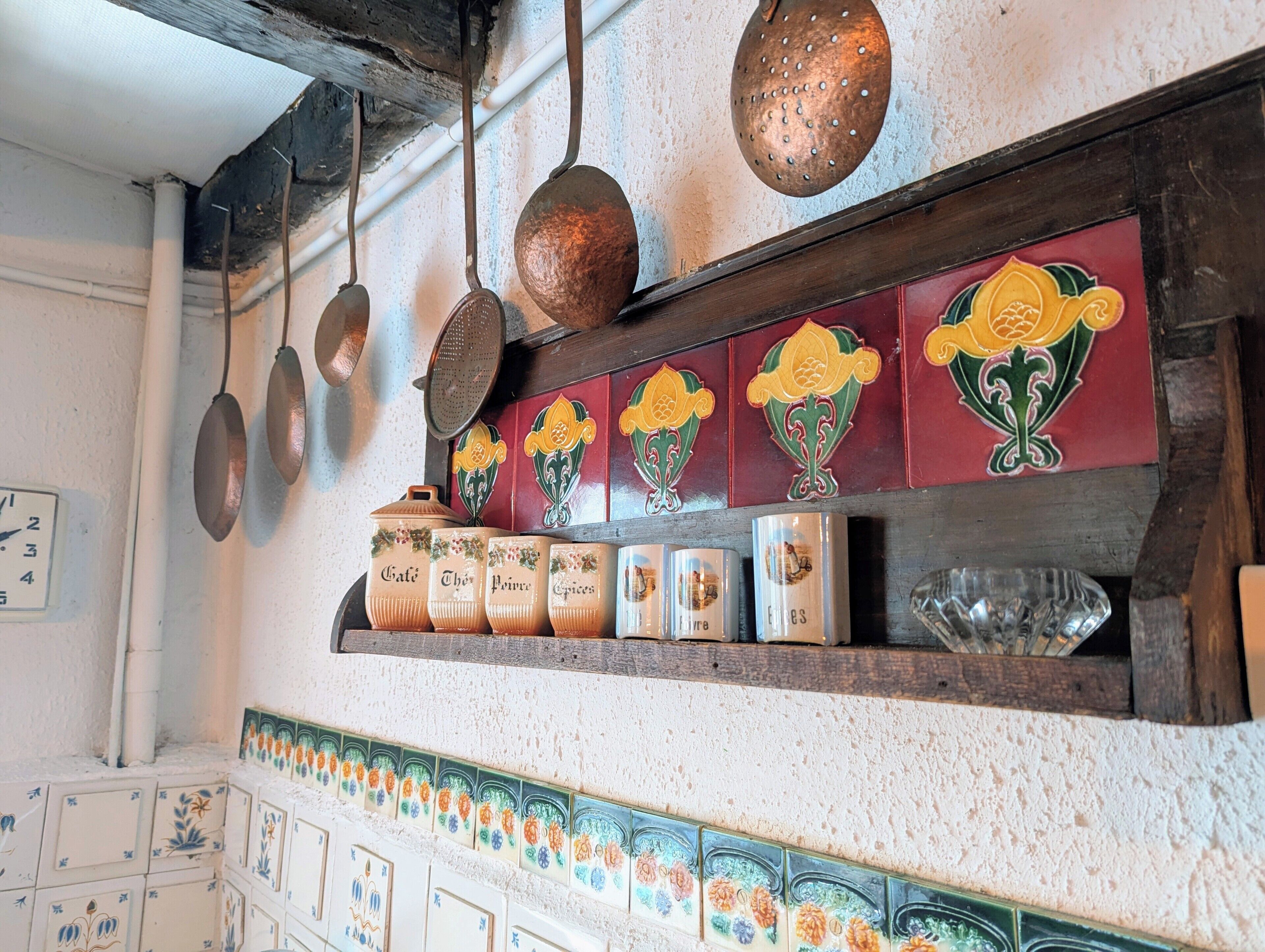 Una cocina con ollas de cobre colgantes, un estante con recipientes de cerámica y un friso decorativo de azulejos.
