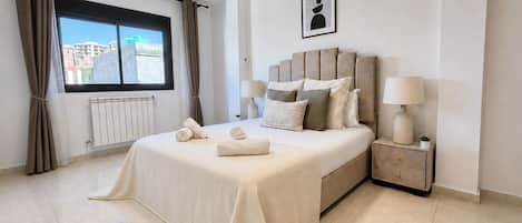 Habitación de hotel con cama, lámparas de noche, radiador, ventana con cortinas e imagen enmarcadas en la pared.