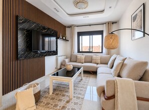 A modern living room with a sectional sofa, a flat-screen TV mounted on a marble wall, a small coffee table, and a large window with curtains.