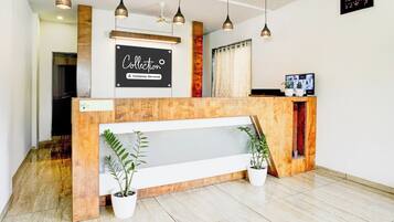 A reception desk with a sign that reads 'Collection Company-Serviced.' There are potted plants on either side of the desk, a digital clock on the wall, and a television screen displaying a cityscape.