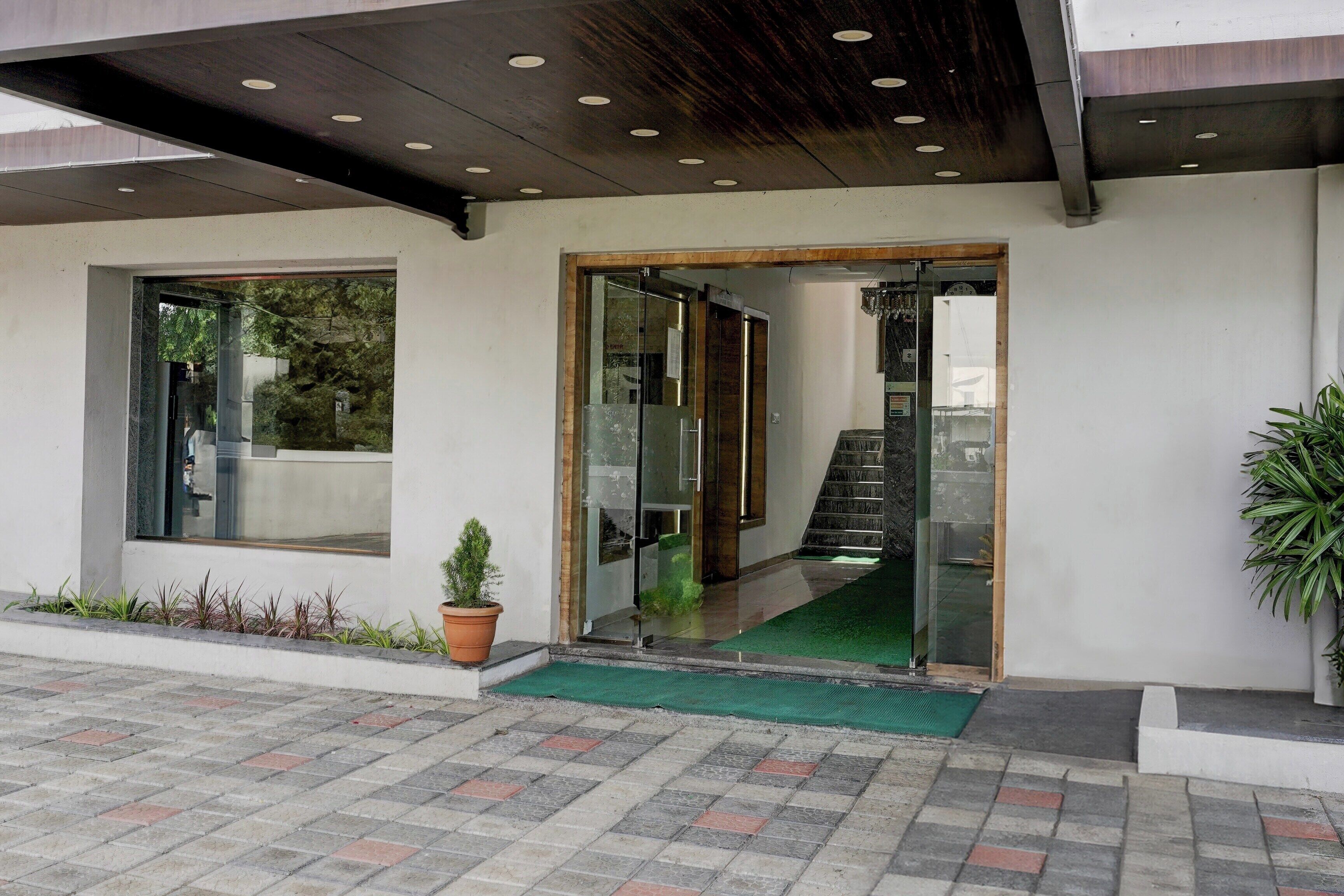 A building entrance with a glass door, a small potted plant, a green mat, and a tiled pavement.
