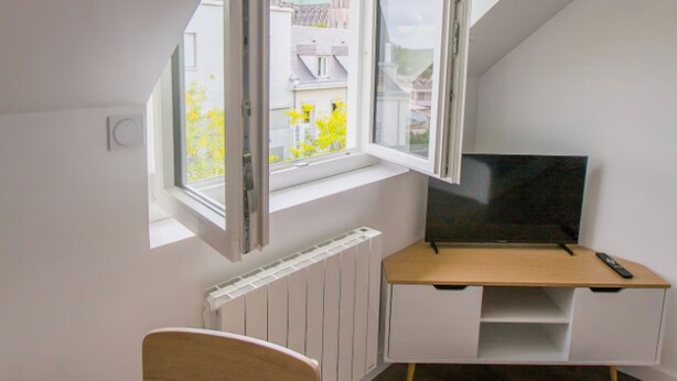 Una habitación con techo inclinado, ventana con vista a un edificio, televisor sobre un soporte y una silla.