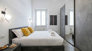 A modern bedroom with a bed, bedside table, and a view into another room. - Italianway Comfort - Cellini 1 (Milan)