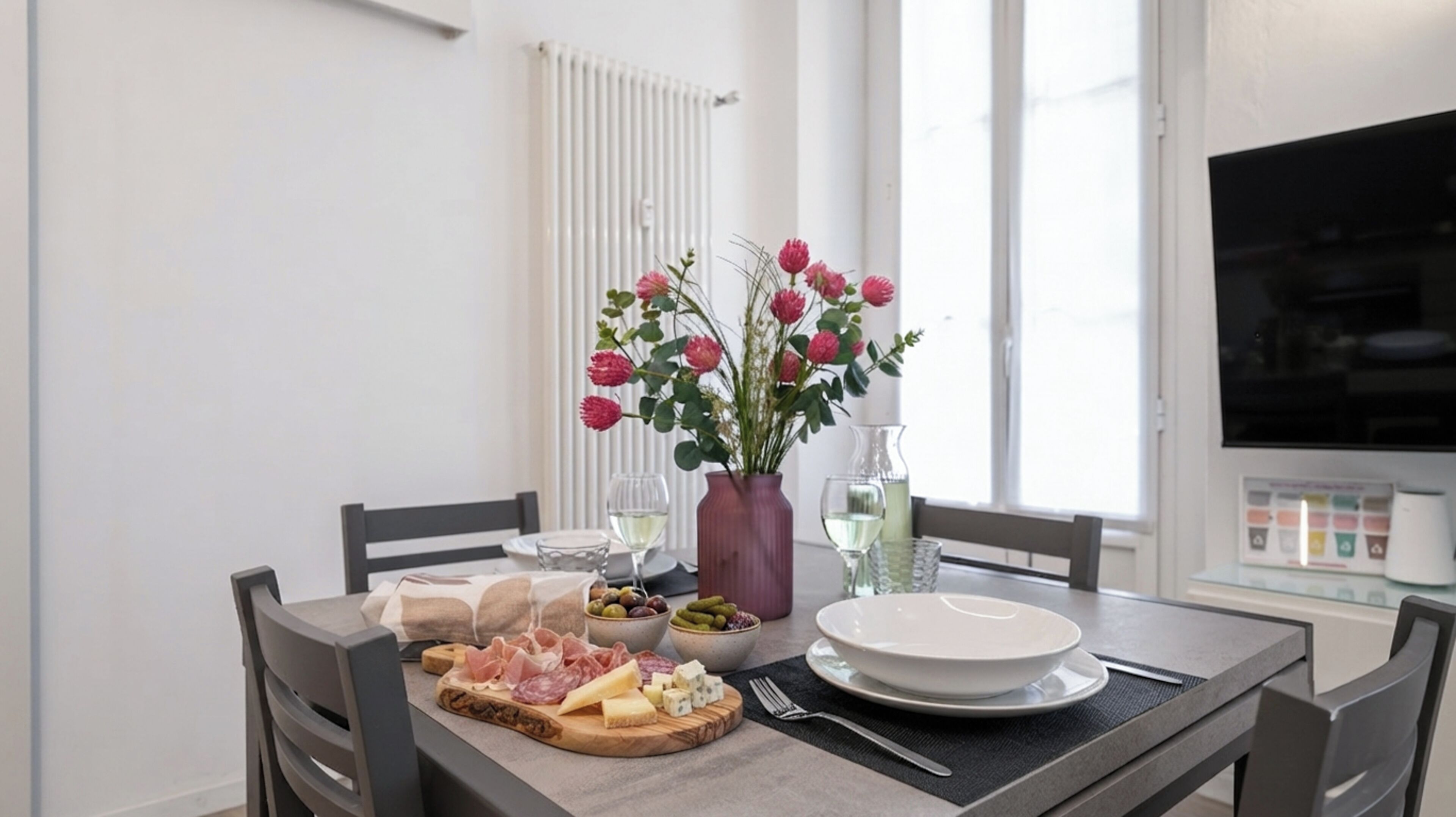 A dining table set with a variety of cheeses, cured meats, olives, and pickles, accompanied by a vase of flowers and wine glasses.