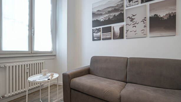 A living room with a sofa, a table with a magazine, a radiator, and a window. - Italianway Comfort - Cellini 1 (Milan)