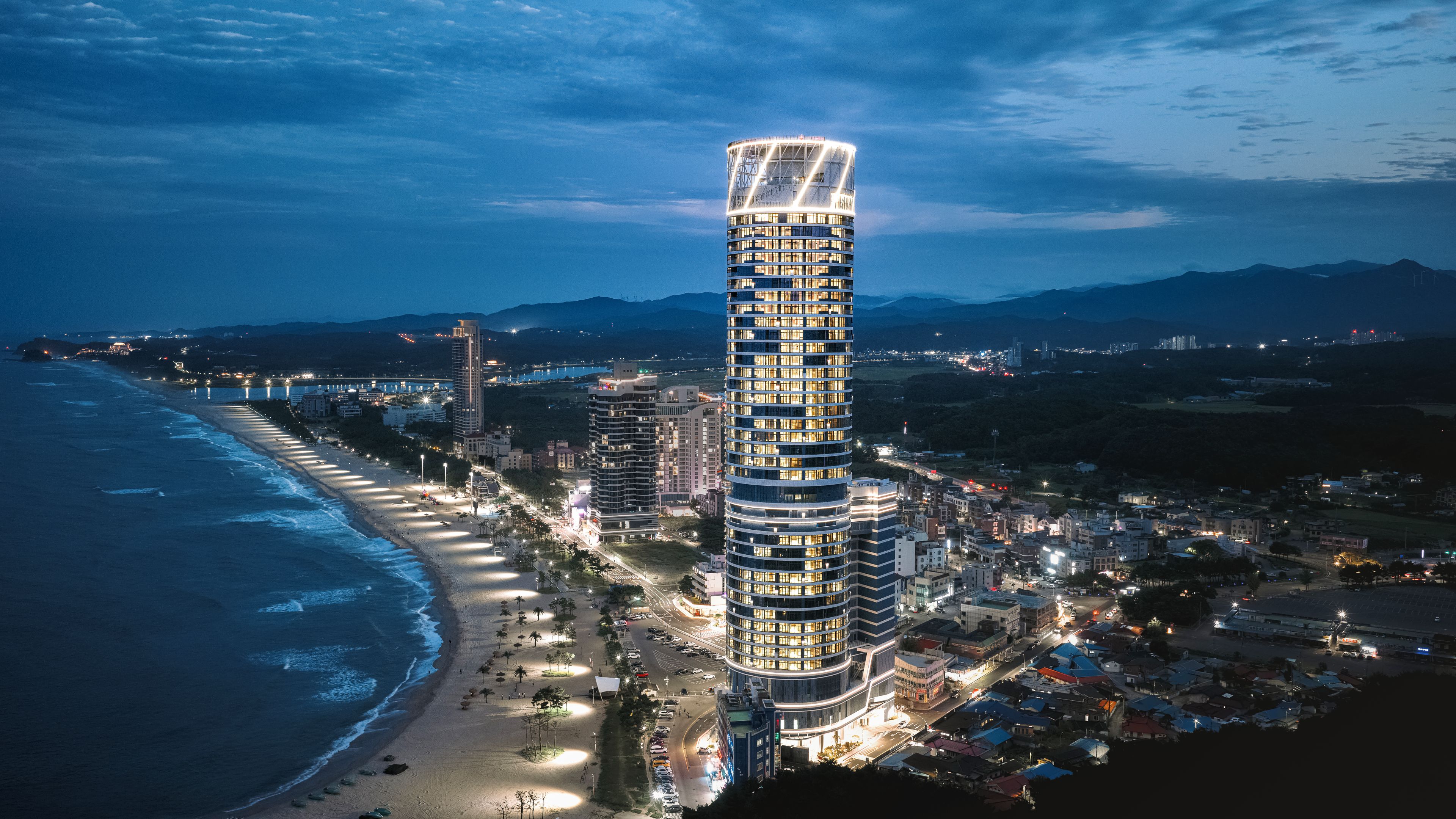 Hotel moderno alto com fachada curva, situado perto de uma praia e calçadão bem iluminado.