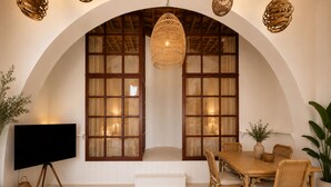 A room with a large arched window, a dining table with chairs, a hanging light fixture, and a television on a tripod. - Villa located in Lindos (Rhodes)
