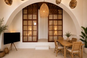 Um quarto com uma grande janela em arco, uma mesa de jantar com cadeiras, um lustre e uma televisão sobre um tripé.