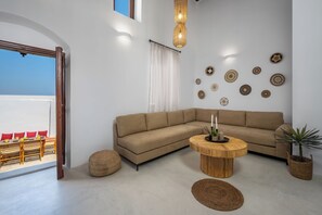 A modern living room with a beige sectional sofa, a round wooden coffee table, and a large window with a view of a dining area. - Villa located in Lindos (Rhodes)