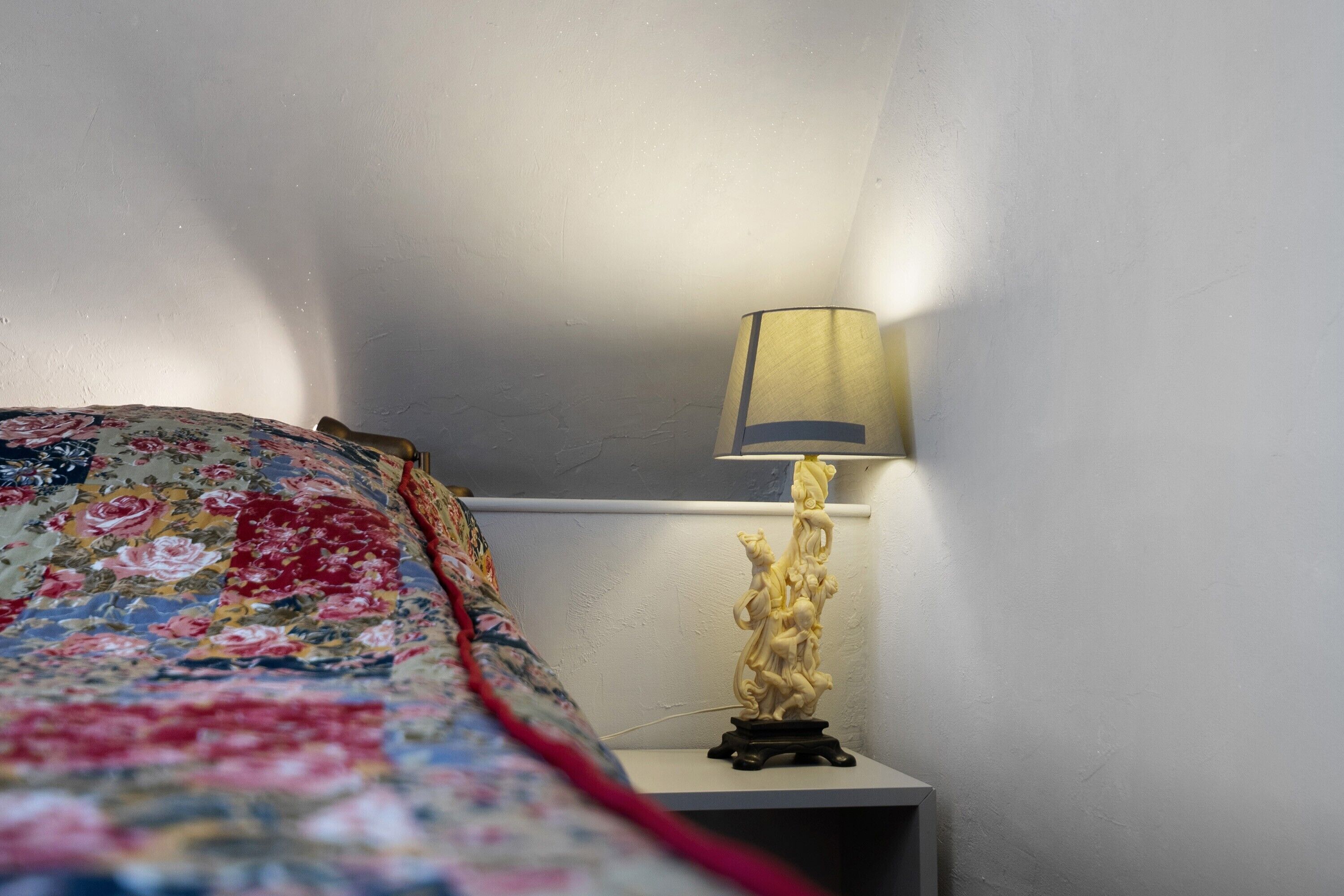 A bed with a floral comforter, a bedside table with a lamp, and a wall.