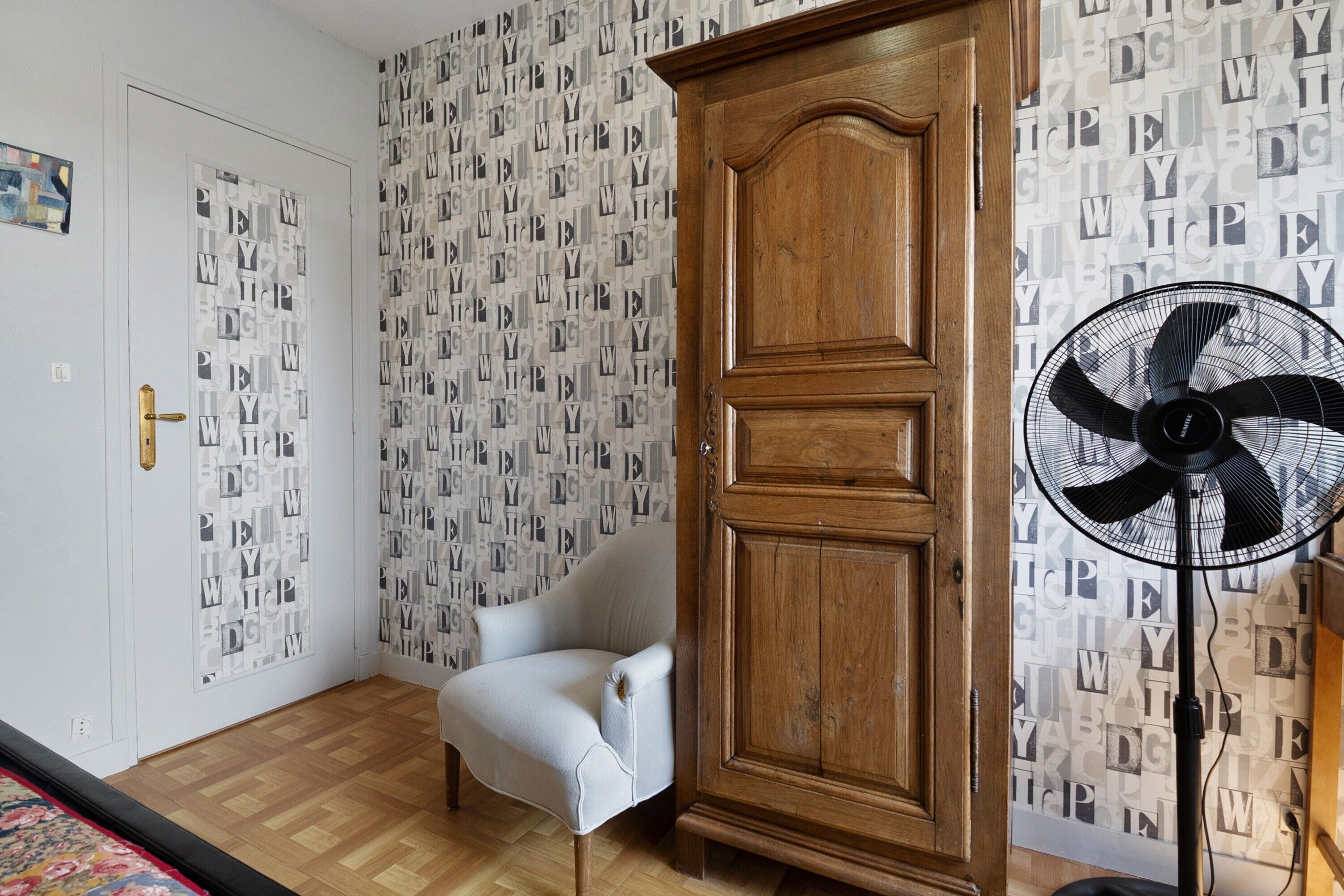 A room with a wooden wardrobe, a white armchair, a patterned wallpaper, and a standing fan.