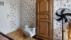 Una habitación con un armario de madera, un sillón blanco, papel tapiz estampado y un ventilador de pie.