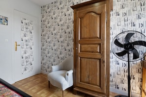 A room with a wooden wardrobe, a white armchair, a patterned wallpaper, and a standing fan.