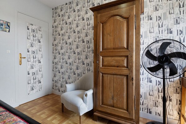 A room with a wooden wardrobe, a white armchair, a patterned wallpaper, and a standing fan.