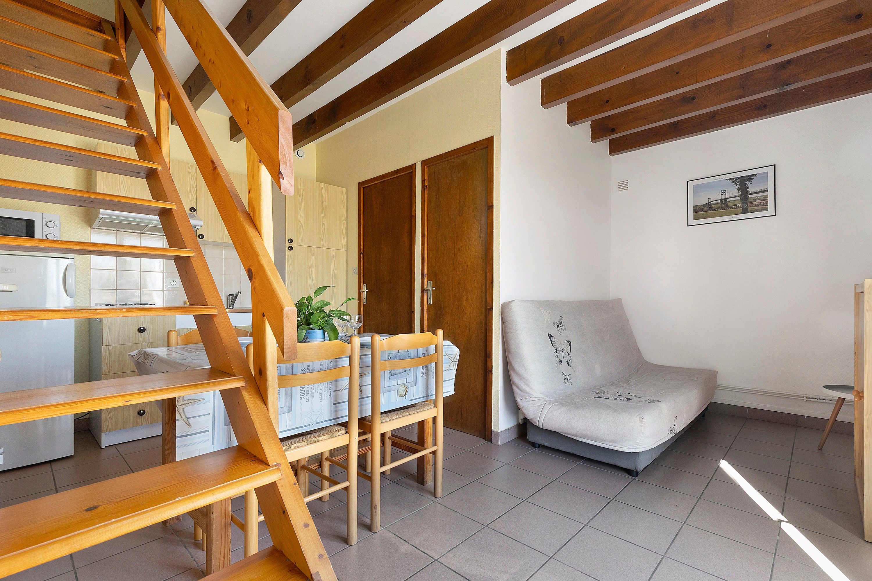 Une pièce avec un escalier en bois, un coin repas avec une table et des chaises, un canapé et un coin cuisine équipé d’appareils électroménagers.