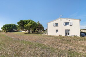 A small, single-story building with a flat roof, a chimney, and a satellite dish. There are two windows with shutters, and a small porch area with outdoor furniture. - Duplex No. 2345 – 40m² – Seaside Residence (Port-des-Barques)
