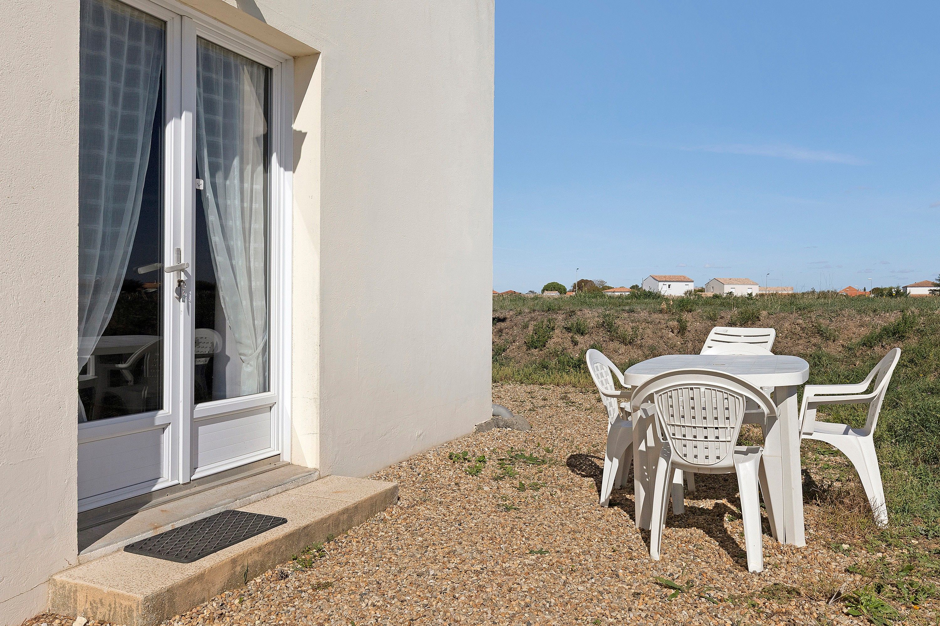 A white building with a door and window, a table with chairs, and a gravel path.