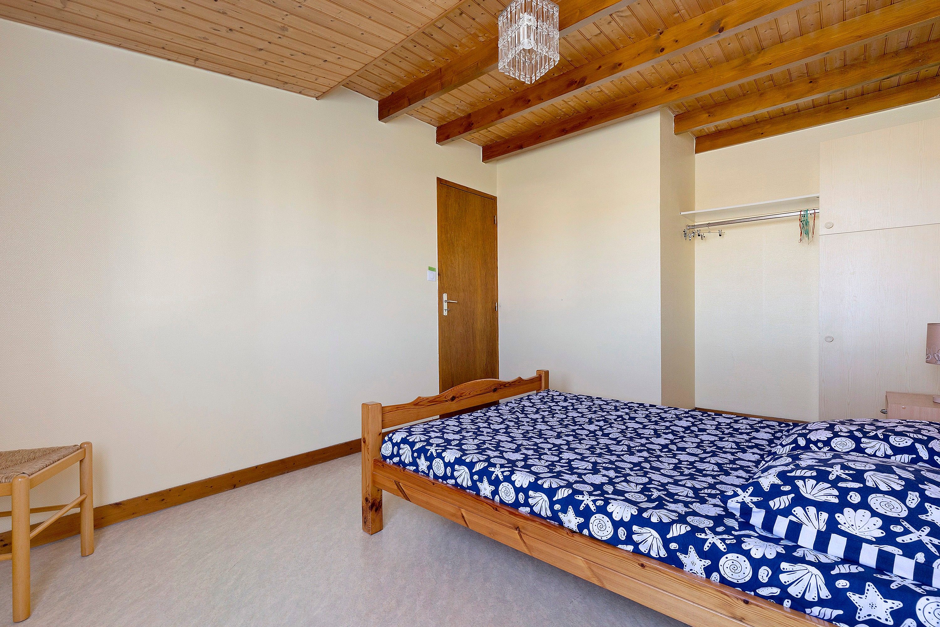 Une pièce avec un lit en bois, une petite chaise en bois, une porte et un plafond avec des poutres en bois et un lustre.
