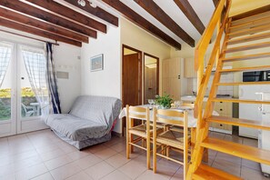 A room with a bed, a dining table with chairs, a wooden staircase, and a kitchen area. - Duplex No. 2345 – 40m² – Seaside Residence (Port-des-Barques)