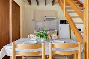A dining area with a table set for two, wooden chairs, a potted plant, and a kitchen in the background. - Duplex No. 2345 – 40m² – Seaside Residence (Port-des-Barques)