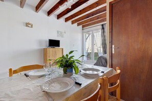 Un coin repas avec une table dressée pour deux personnes, un téléviseur posé sur un meuble en bois et une fenêtre avec des rideaux.
