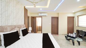 A hotel room with a bed, a ceiling fan, an air conditioning unit, and a glass table with a kettle and cups.