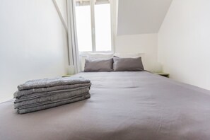 A neatly made bed with a grey bedspread and pillows, a stack of folded towels, and a window with curtains. - Appartement le Off (Chartres)
