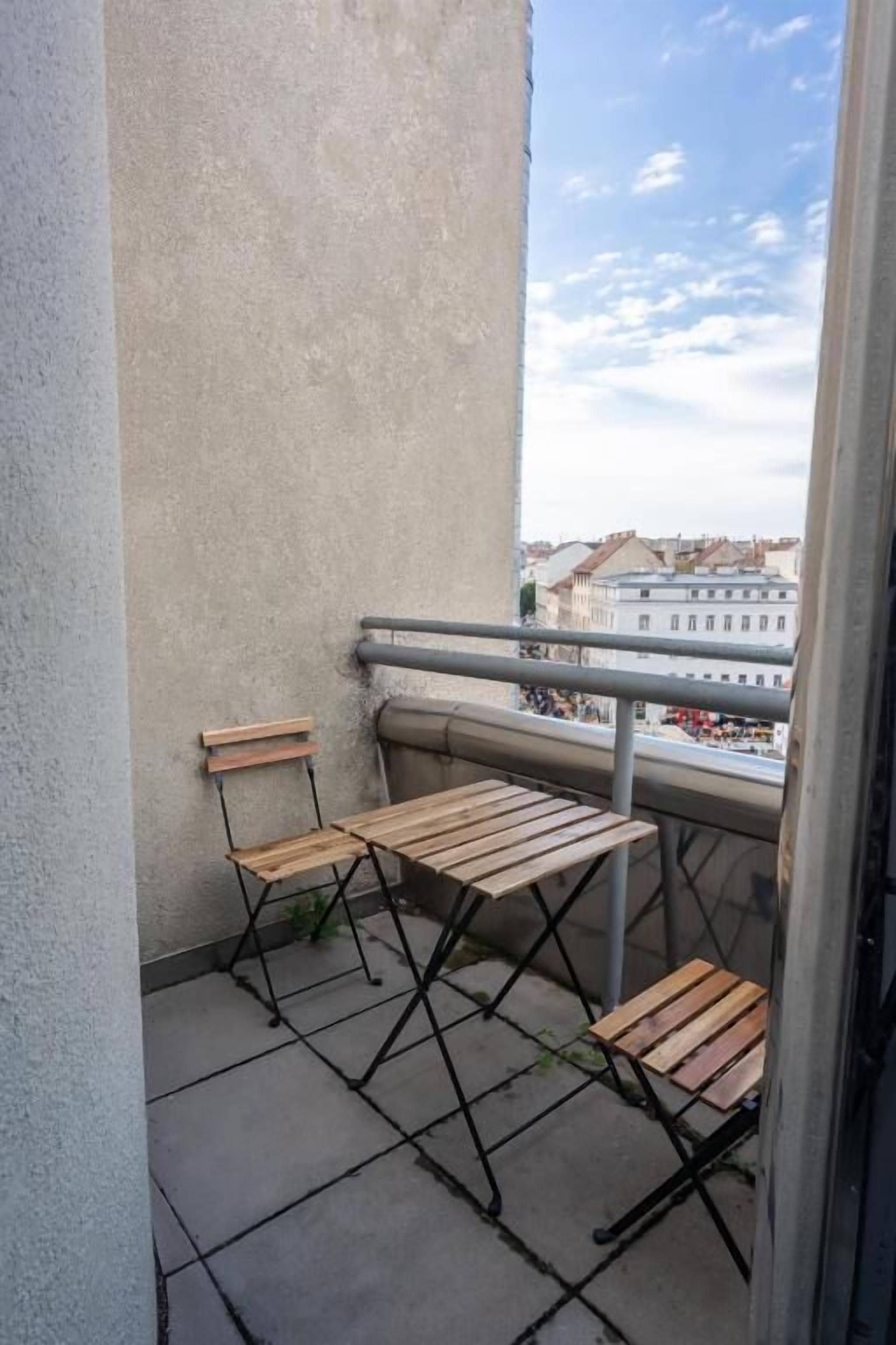 Un balcón con dos mesas y sillas de madera, barandilla metálica y vistas a edificios en la distancia.