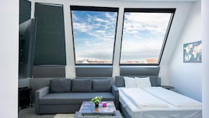 Un dormitorio moderno con cama, sofá, mesa de centro y vistas a la ciudad a través de amplios ventanales.