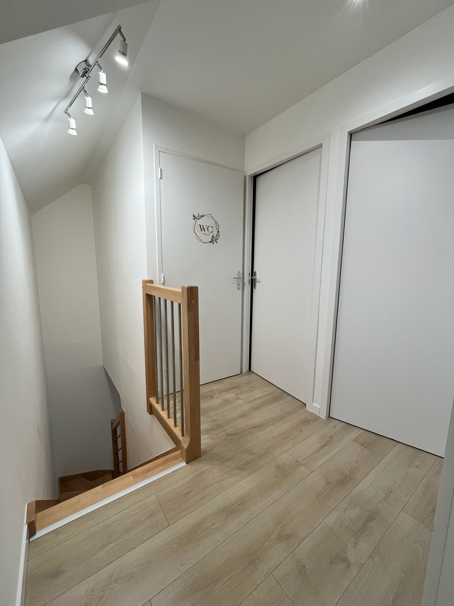 A modern interior with a wooden staircase, a white door with a 'WC' sign, and a light-colored wooden floor.