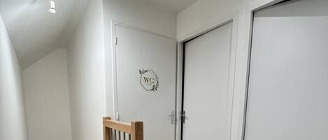 Un interior moderno con una escalera de madera, una puerta blanca con un letrero que indica 'WC' y un suelo de madera claro.