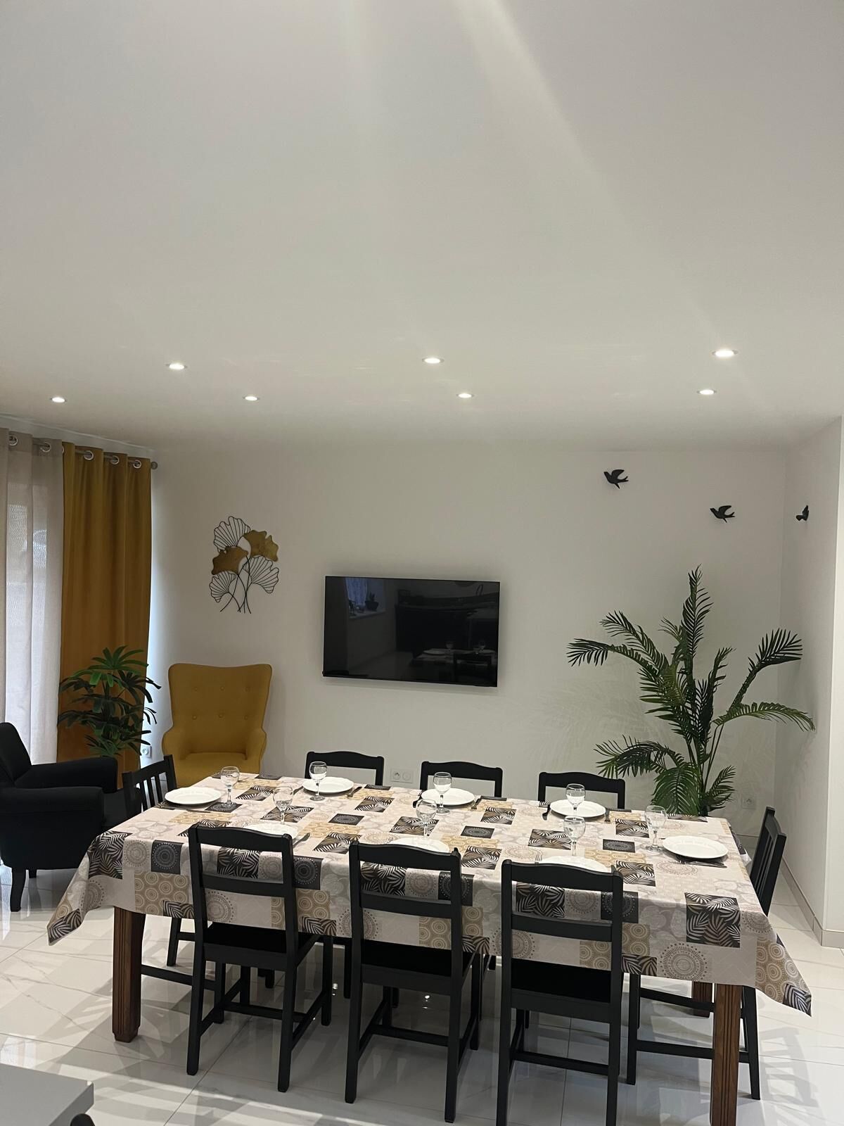 Un comedor con una mesa dispuesta para seis personas, rodeada de sillas, un televisor montado en la pared y decoración artística en la pared.