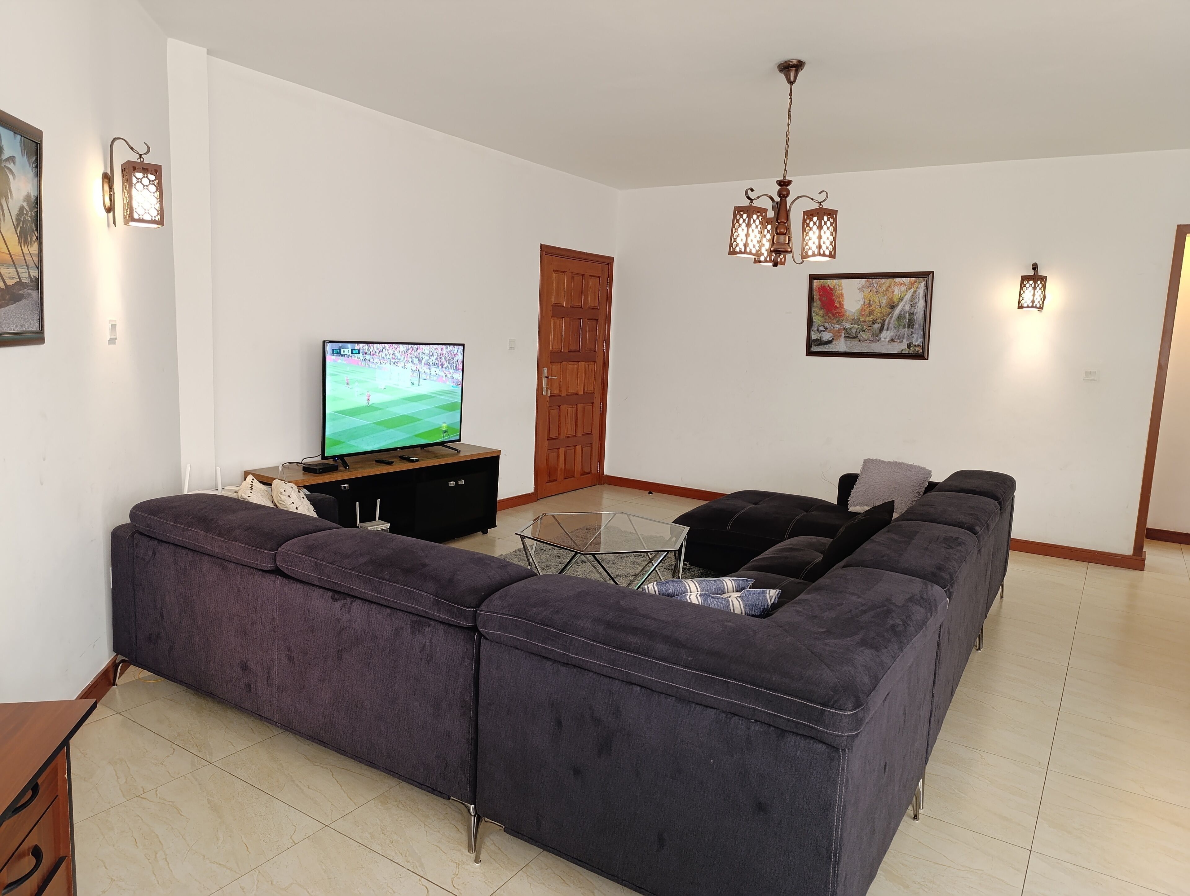 A living room with a large flat-screen TV, a dark-colored sofa, a glass coffee table, and a wooden door.