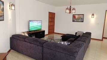 A living room with a large flat-screen TV, a dark-colored sofa, a glass coffee table, and a wooden door.