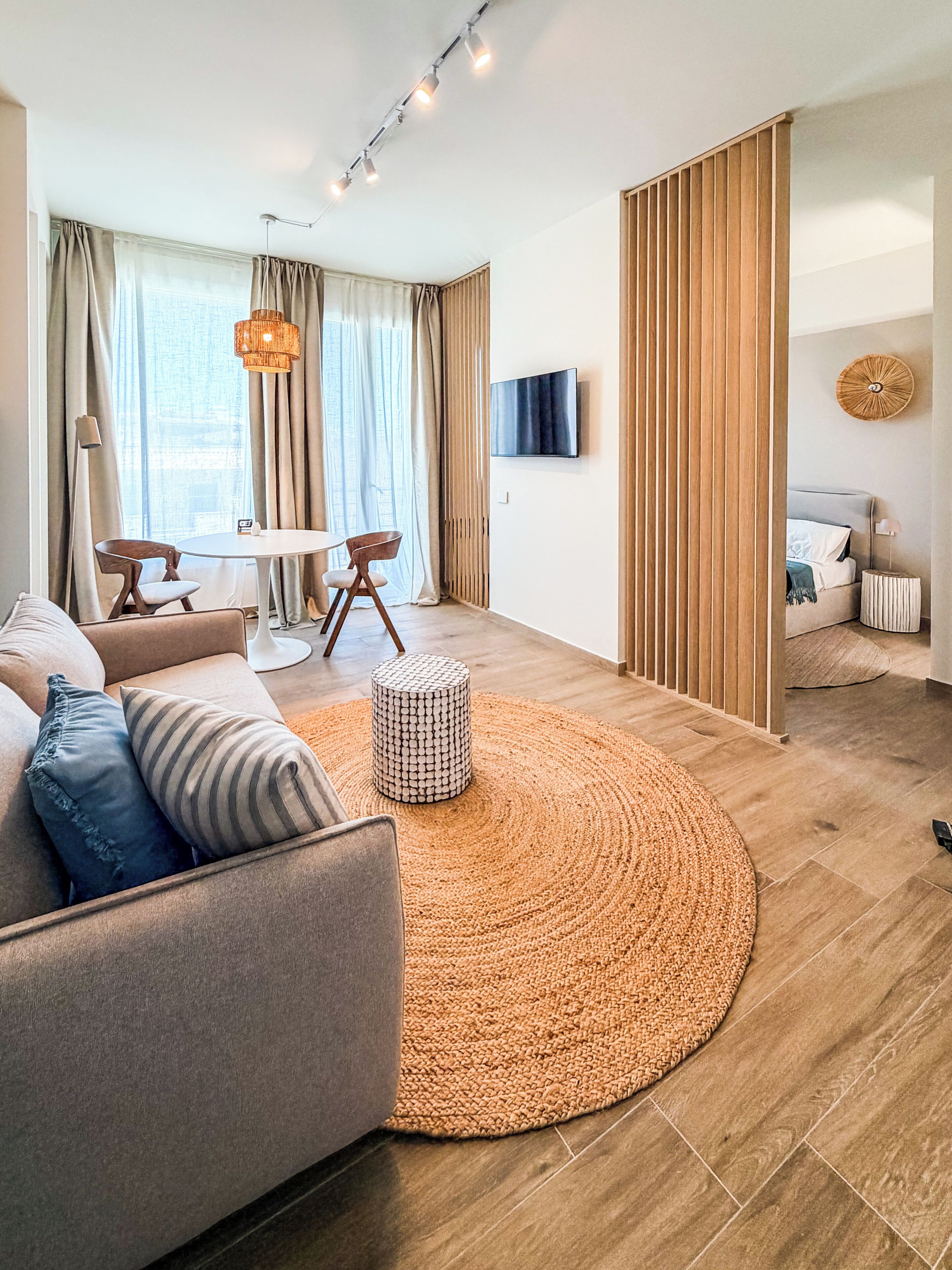 Een moderne woonkamer met een bank, een rond tapijt, een eettafel met stoelen en een aan de muur bevestigde televisie.