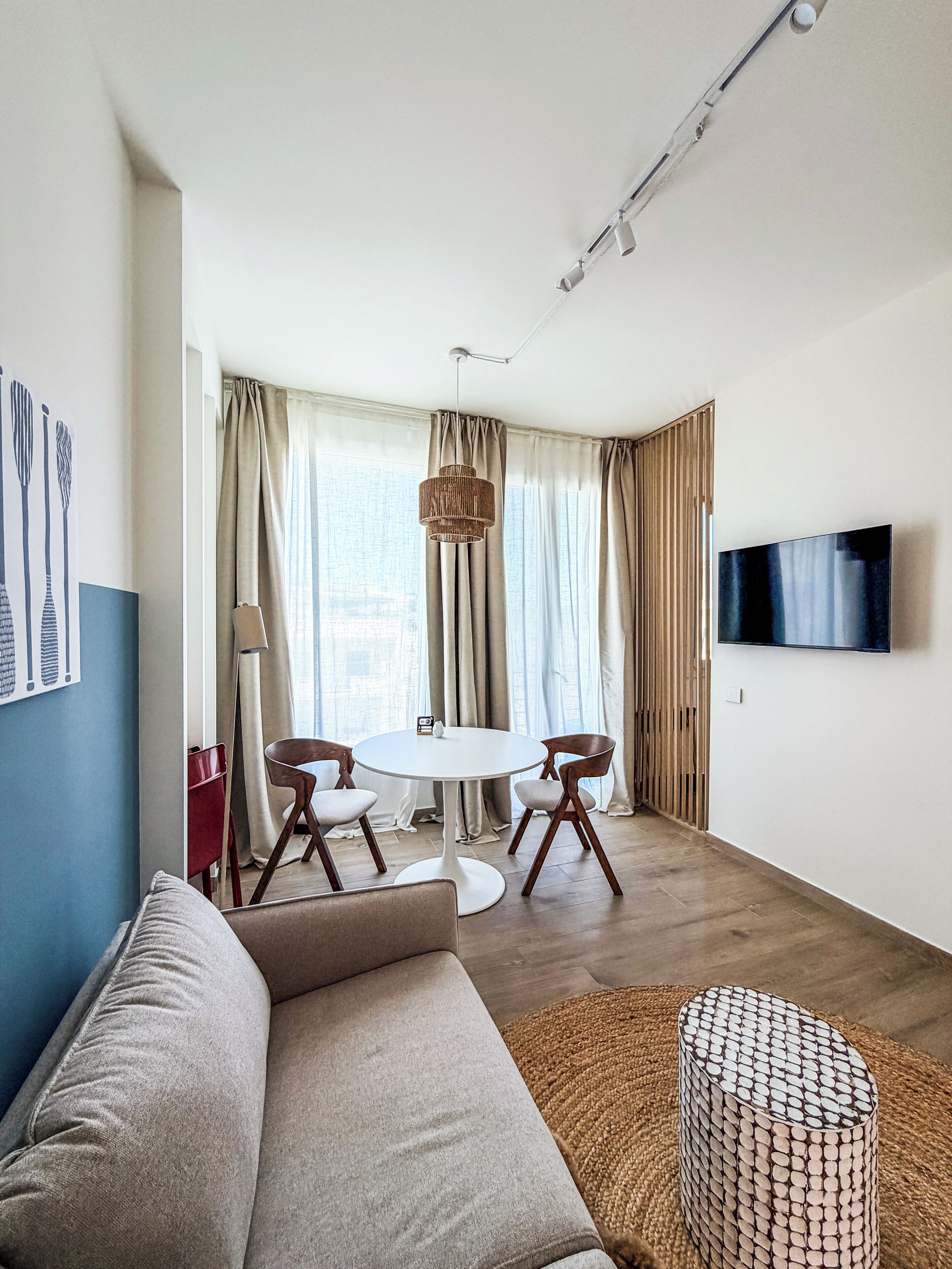 A modern living room with a round dining table, two chairs, a sofa, a TV mounted on the wall, and a window with curtains.
