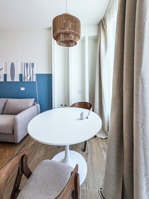 Ein modernes Wohnzimmer mit einem runden weißen Tisch, einem grauen Sofa, einem Holzstuhl, einer Weidenpendelleuchte und Bildern an der Wand.