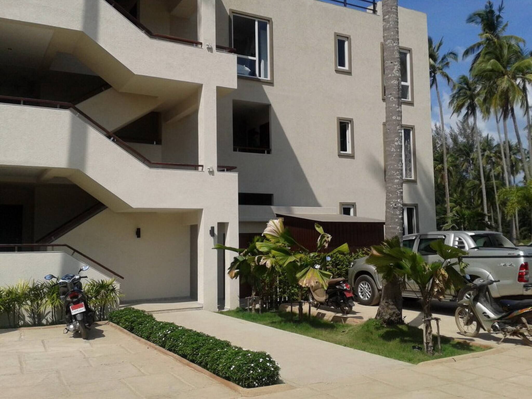 A multi-story building with balconies, a parking area, and a small garden with tropical plants.