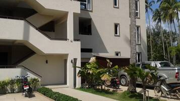 A multi-story building with balconies, a parking area, and a small garden with tropical plants.
